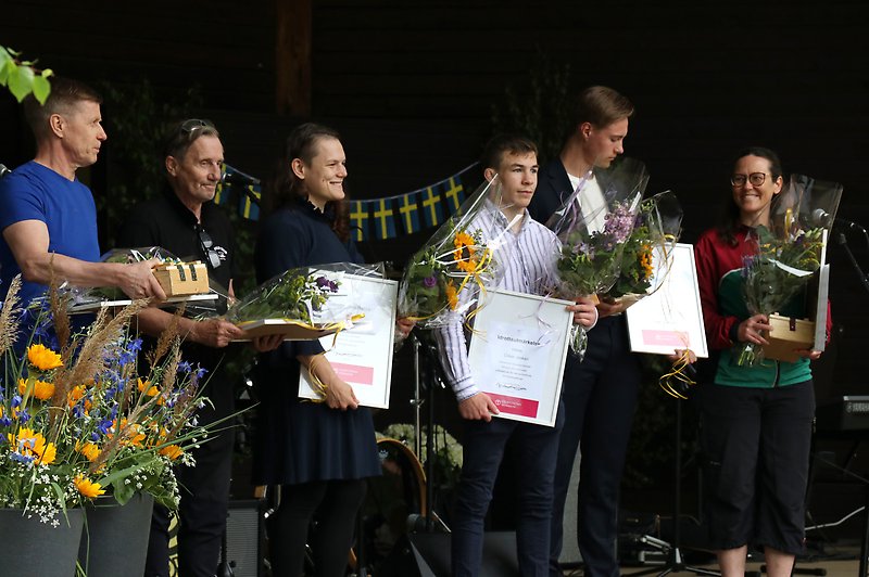 Flera glada vinnare blickar ner från scenen i Holje park med blommor och diplom i famnen. Foto.